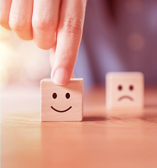 Une main tient entre le pouce et l’index un cube en bois posé sur une table. Sur la face du cube exposé à la caméra se trouve un bonhomme sourire. En arrière-plan, on voit de manière floue un autre cube qui expose un bonhomme triste.