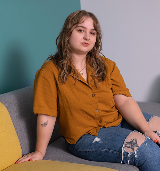 Une jeune femme est assise sur un sofa et fixe la caméra. Son jeans déchiré à la hauteur du genou laisse voir un tatouage.