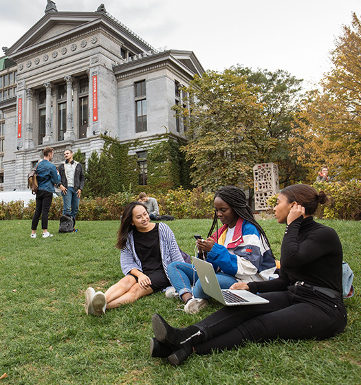 Groupe diversifié d'étudiants socialisant sur l'herbe du campus inférieur de McGill avec le musée Redpath en arrière-plan.